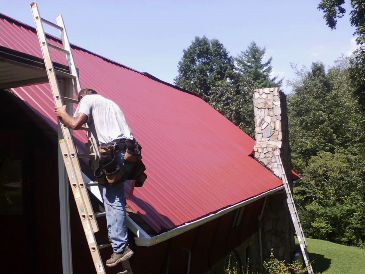 Roofer on a ladder beside a red metal roof and stone chimney — Soffit & Fascia Repair by Roofing Brothers in Erath